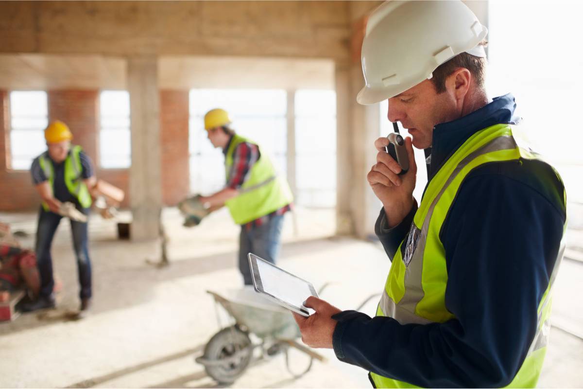 Construction site supervisor with tablet and radio, workers in background.
