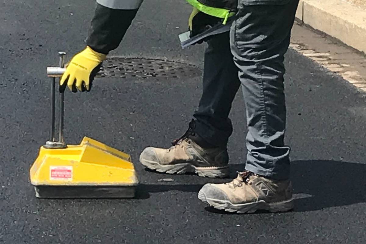 Worker operating a yellow nuclear density gauge on new asphalt pavement.